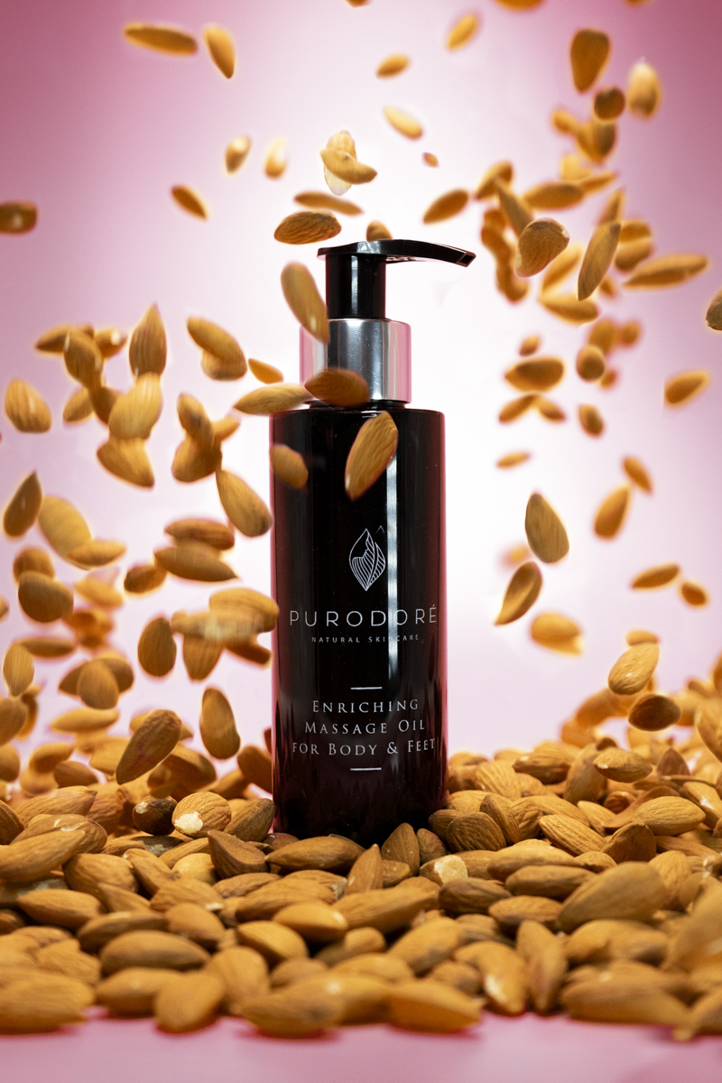 Black bottle of PURODORÉ natural skincare enriching massage oil for body and feet surrounded by and falling almonds on a pink background.