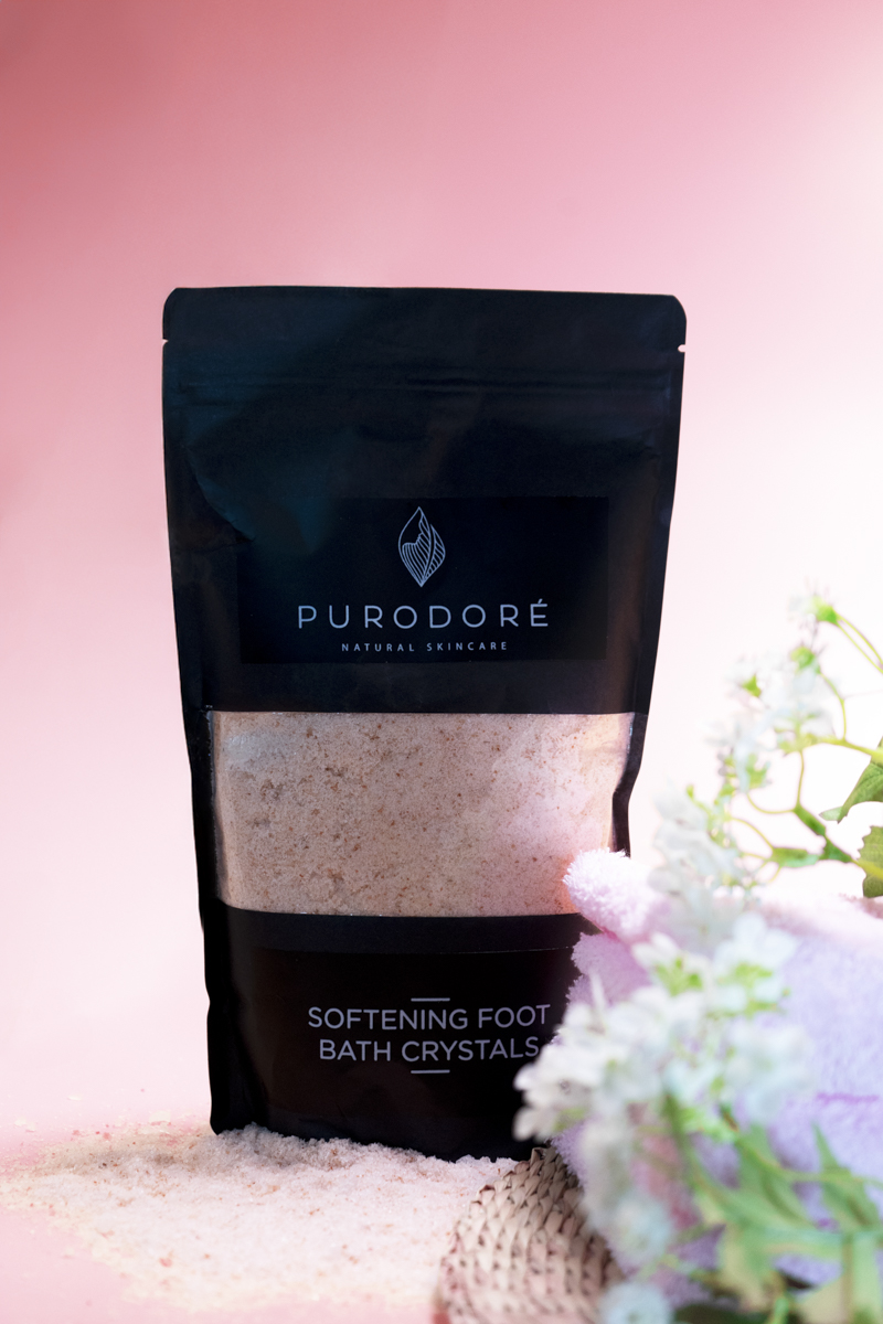 Black resealable pouch of Purodoré Natural Skincare softening foot bath crystals, with pinkish crystals spilled at the base, set against a pink background with white flowers and a pink towel nearby.