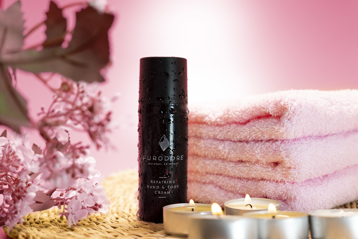 Black bottle of Purodore repairing hand and foot cream with water droplets, next to three folded pink towels, lit tealight candles, and pink flowers on a wicker surface with a pink background.
