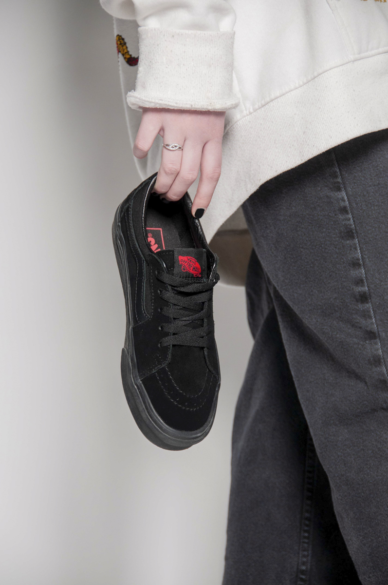 Person wearing a white sweatshirt and dark pants holding a black Vans sneaker by the heel with their left hand, showing the shoe's red Vans Off The Wall logo on the tongue.