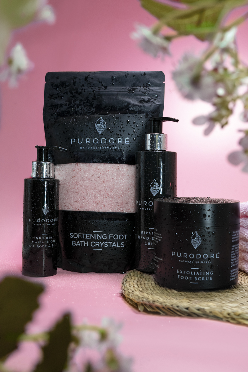 Four Purodoré natural skincare products against a pink background: softening foot bath crystals in a black pouch, enriching massage oil in a pump bottle, repair hand and foot cream in a pump bottle, and exfoliating foot scrub in a jar, all with water droplets on them.