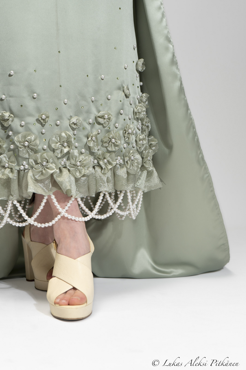 Close-up of a light green dress hem decorated with fabric flowers, pearls, and beads, worn with cream peep-toe high-heeled shoes.