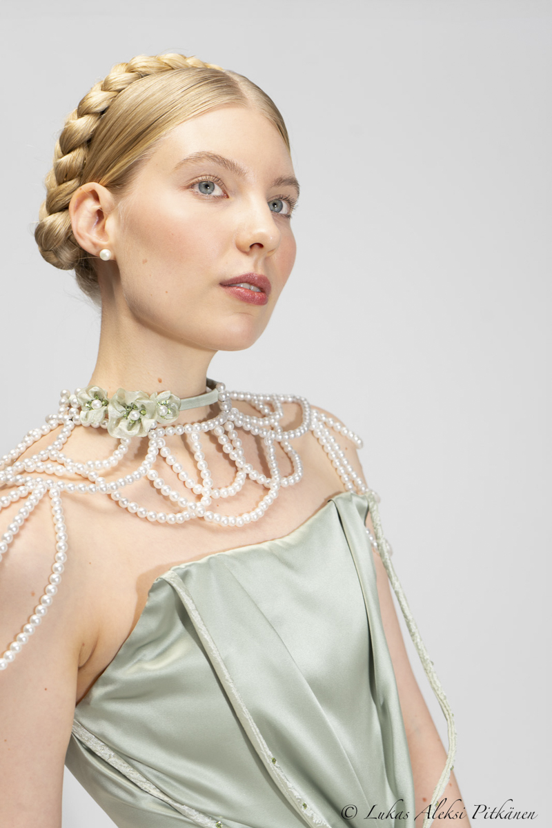 Portrait of a woman with blonde hair styled in a braided crown, wearing pearl earrings and an elegant light green satin dress adorned with a pearl necklace and floral choker.