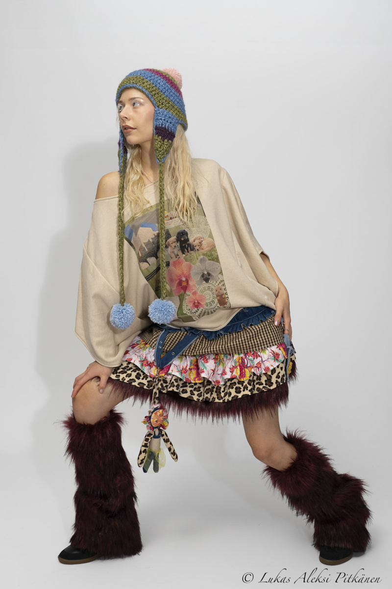Young woman posing against a plain background wearing a beige off-shoulder top with a puppy and flower print, a multi-patterned layered skirt with leopard print and floral ruffles, burgundy furry leg warmers, and a colorful knit hat with ear flaps and blue pom-poms.