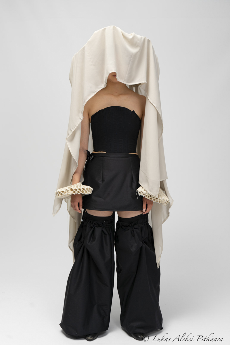 Person standing against a plain light background wearing an avant-garde outfit with a black strapless corset top, a short black skirt, oversized black wide-leg pants, and a light beige fabric draped over the head and shoulders, partially covering the face and arms.