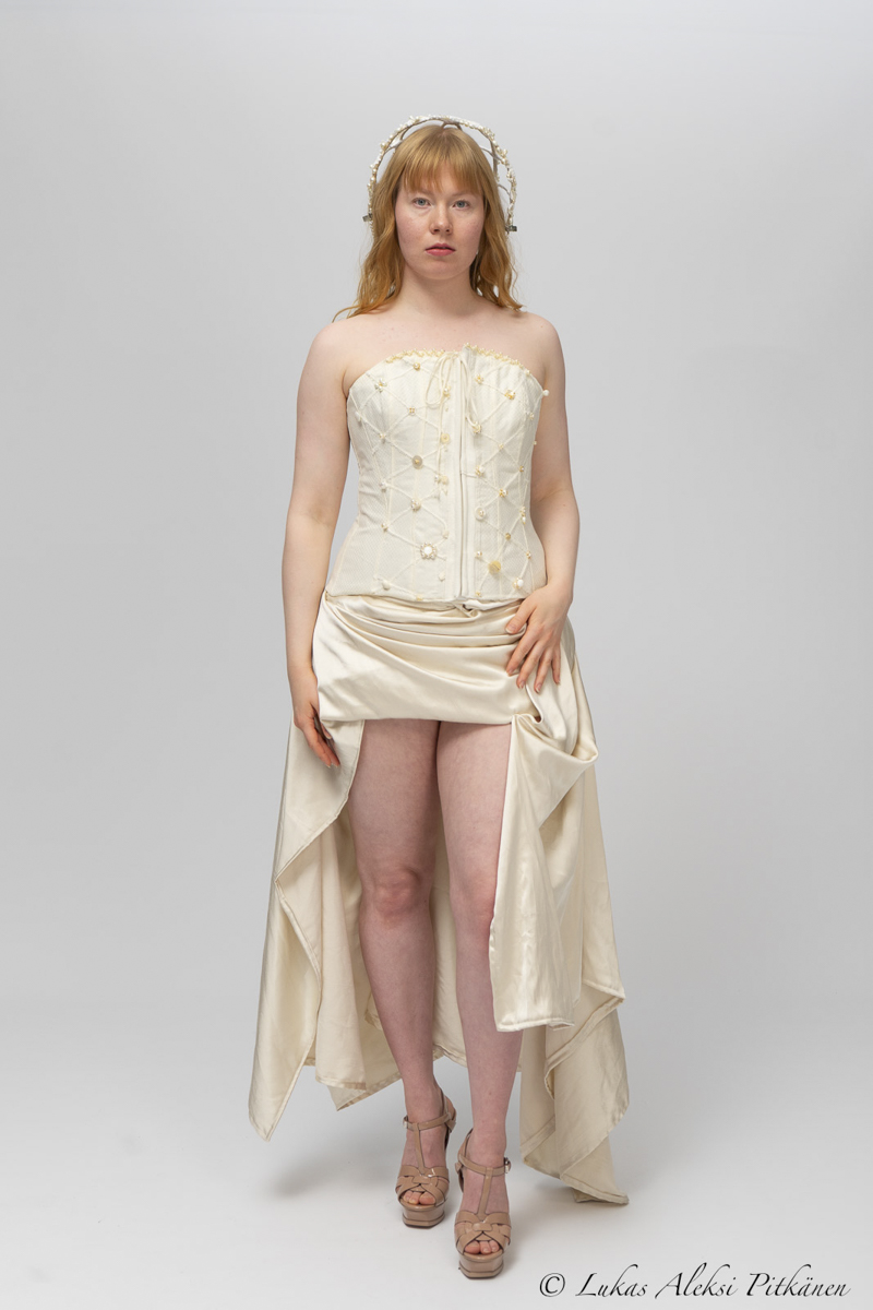 Woman with light skin and red hair wearing a cream-colored strapless corset with floral embroidery, a draped satin high-low skirt, beige platform heels, and a matching headband, standing against a plain light gray background.