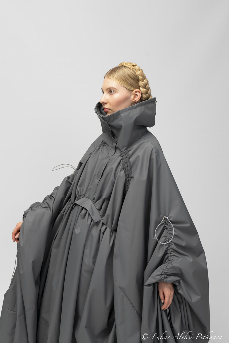 Side view of a woman with blonde braided hair wearing a voluminous gray rain poncho with a high collar and gathered elastic details on the sleeves and chest.