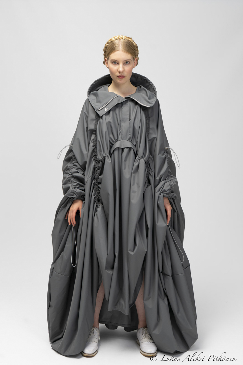 Woman with braided hair wearing a loose, oversized gray dress with gathered, voluminous fabric and wide sleeves, standing against a plain gray background, wearing white shoes.
