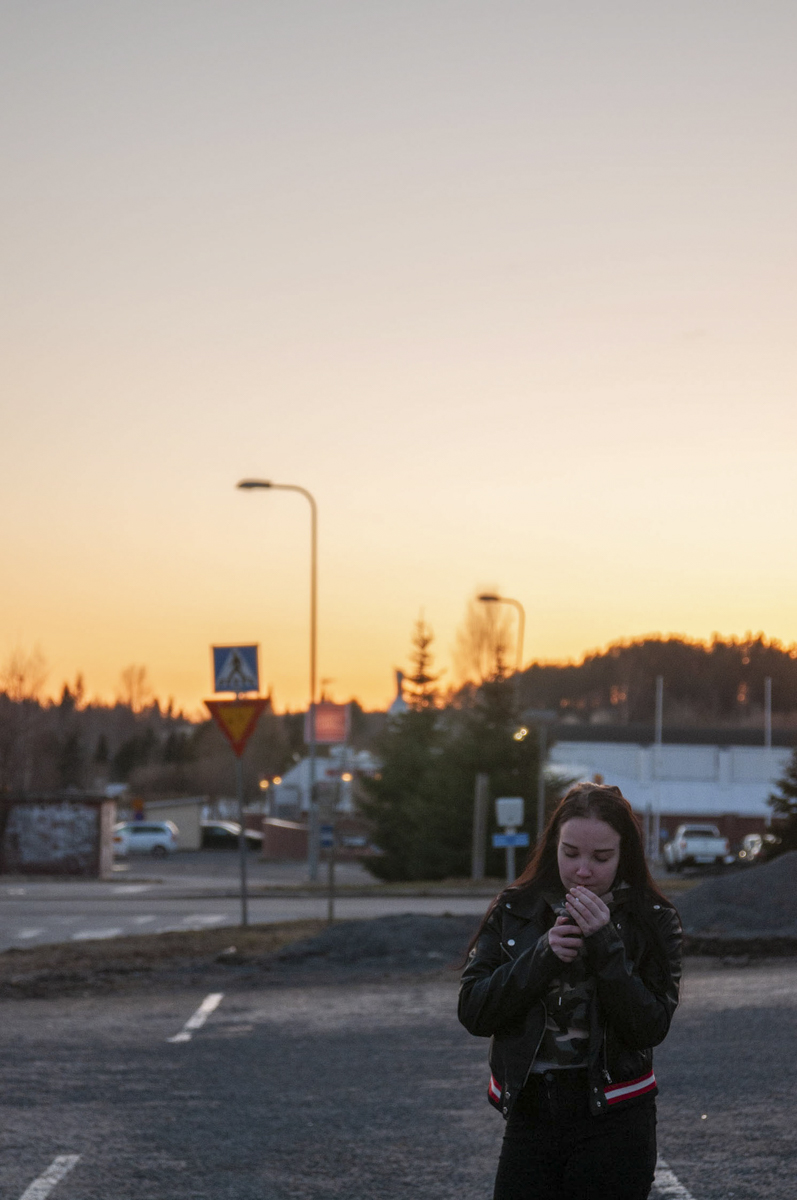 Young woman wearing a black jacket stands alone on a parking lot at sunset, looking down at her hands as the sky glows orange behind her.