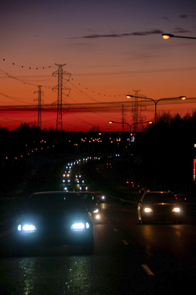 Cars with headlights on driving on a road at dusk under a vibrant orange and purple sky, with streetlights and power lines visible.