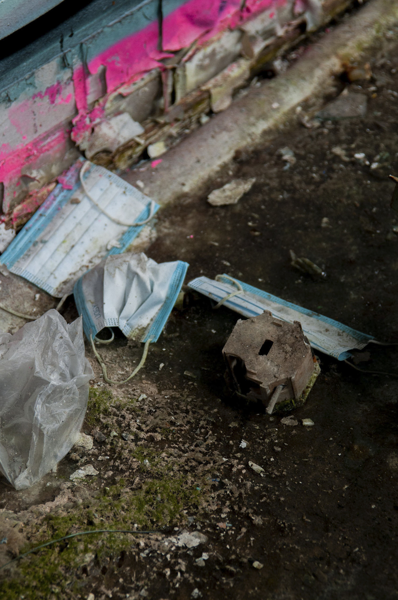 Discarded dirty blue and white face masks, a crumpled plastic glove, and a rusted metal piece on a grimy floor with chipped paint and green moss near a wall painted with peeling teal and pink layers.