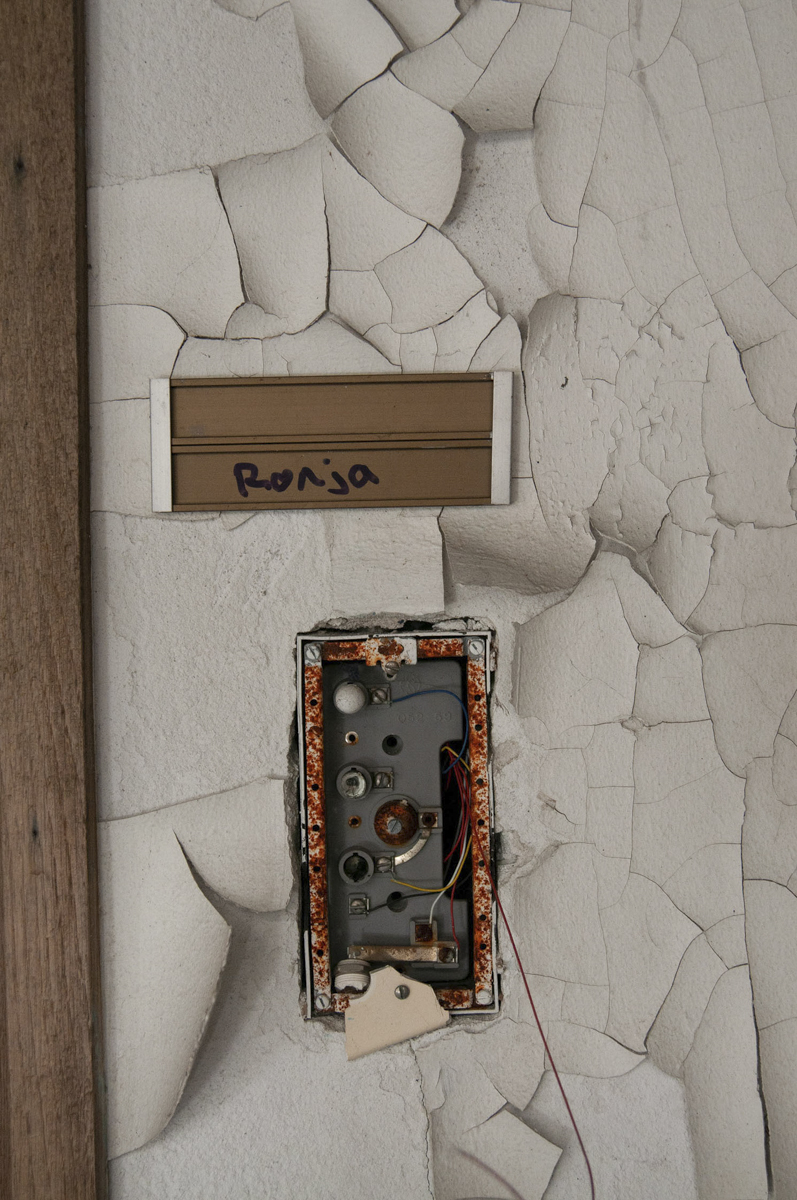 Close-up of a cracked and peeling white wall with an empty, rusted metal electrical box and exposed wires, below a nameplate labeled 'Ronja' in black ink on a brown background.