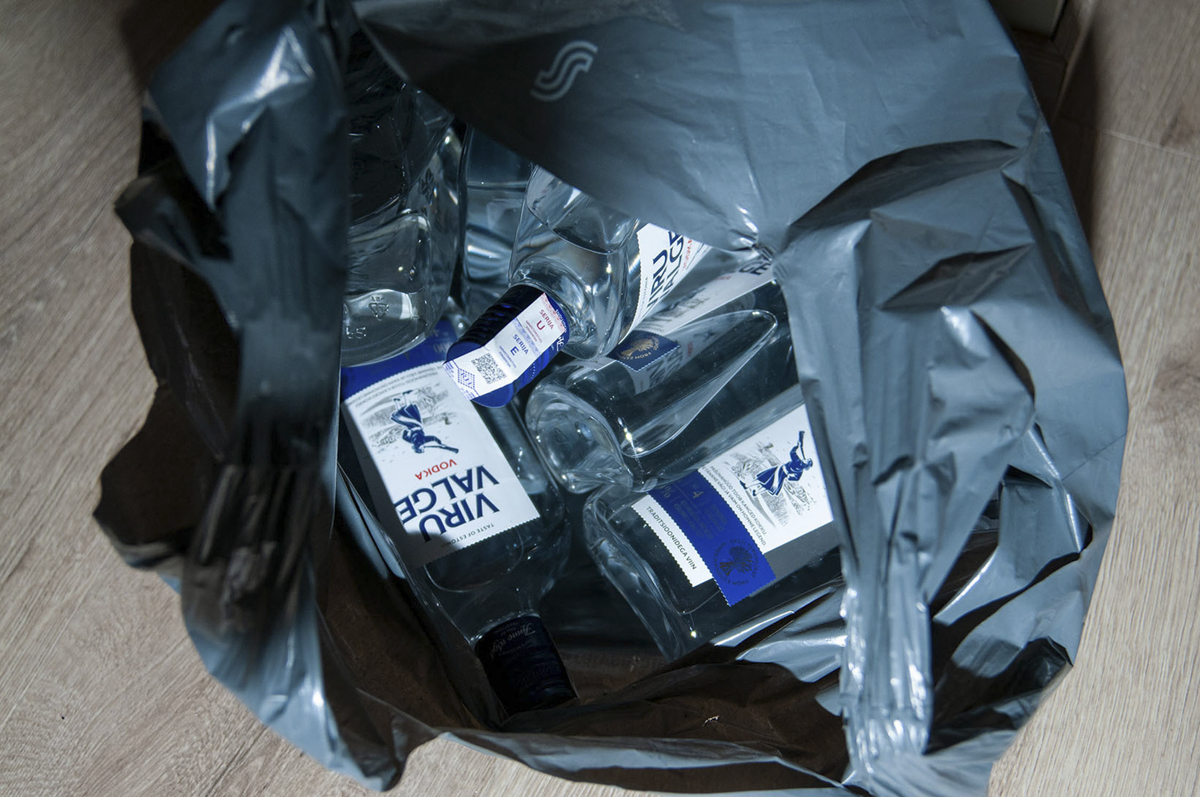 Several empty vodka bottles with blue and white labels inside a black trash bag on a wooden floor.