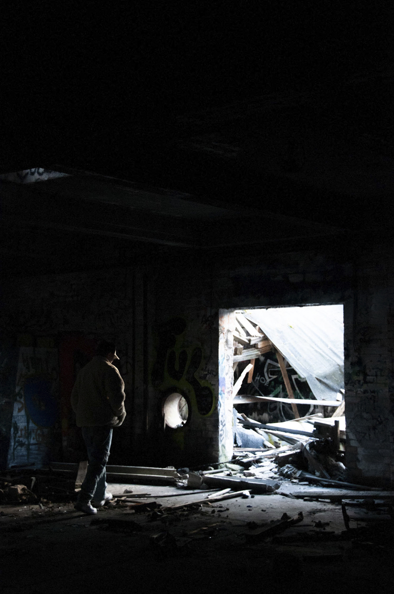 A person in a light jacket and jeans stands in a dark, abandoned building near a large opening filled with debris and broken wooden planks, with graffiti visible on the walls.