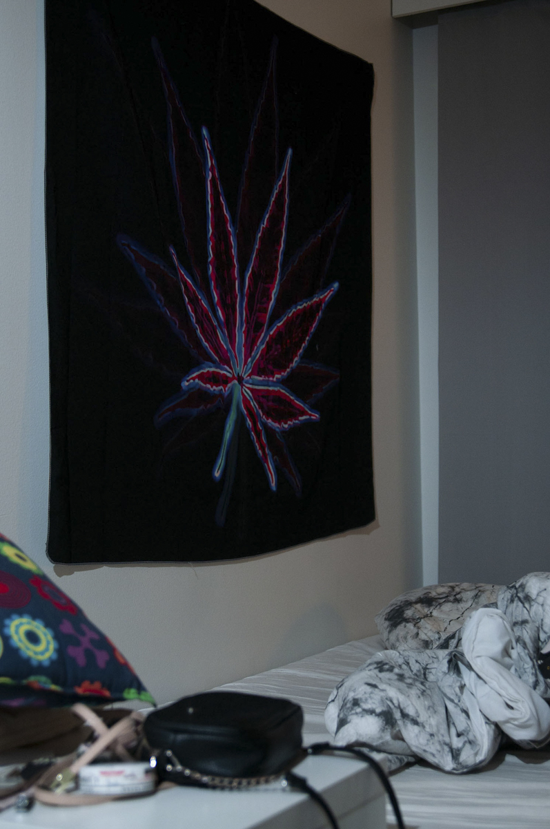 Bedroom corner with a dark tapestry featuring a glowing red and blue stylized leaf hanging on the beige wall, a colorful pillow on the left, a black pouch on a white bedside table, and a crumpled white and gray blanket on the bed.