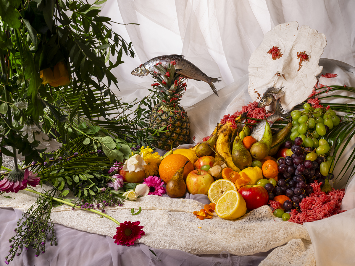 A still life arrangement of various fruits including bananas, pears, apples, grapes, oranges, and lemons on fabric with flowers and green leaves; two whole fish are placed among the fruits, one balanced on a pineapple and the other near a white clay mask with red floral decorations resembling a face.
