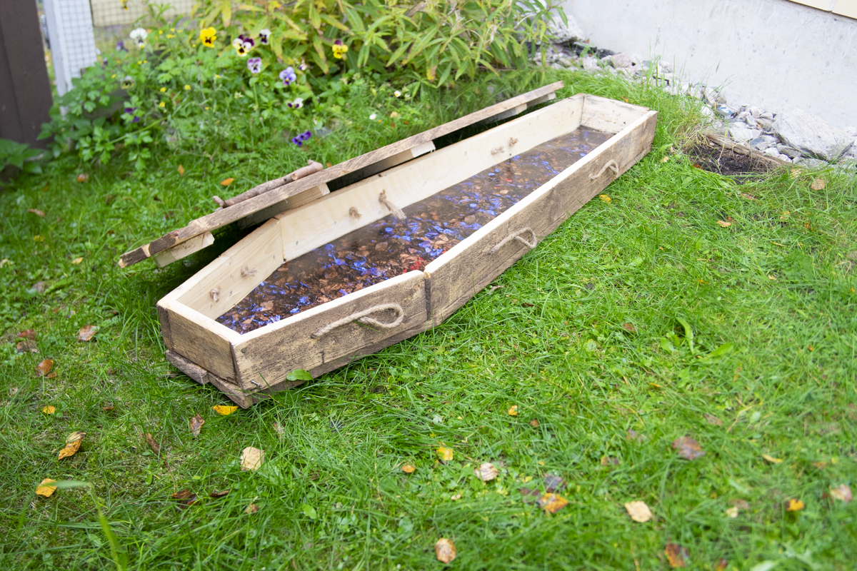 Open rustic wooden coffin with rope handles placed on green grass near a flower bed with yellow and purple flowers.