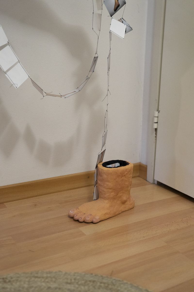Sculpture resembling a large human foot and lower leg made of an orange material sitting on a wooden floor near a white wall and door, with a hanging wire mobile displaying small rectangular photos above it casting shadows on the wall.