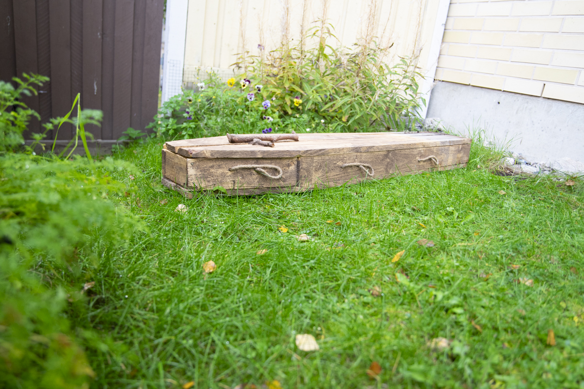 Old wooden coffin with rope handles resting on green grass in a garden area near a fence and a building.