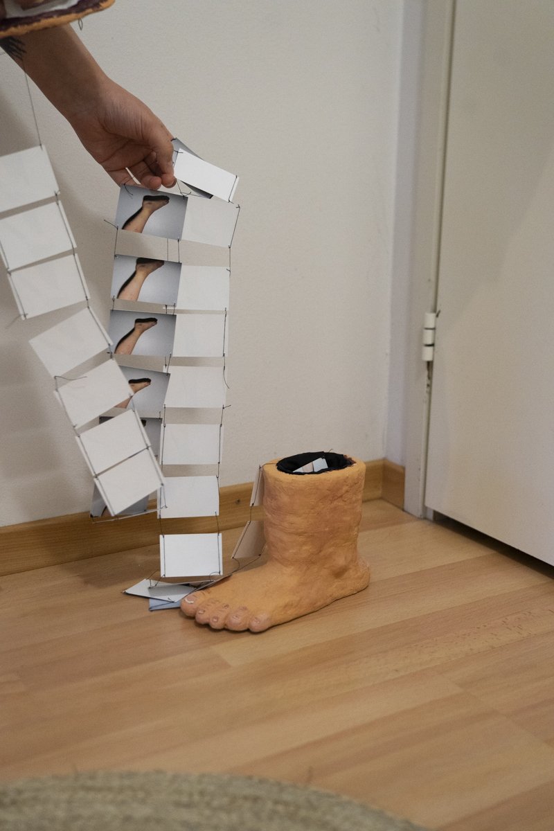 A hand holding a string of small paper frames printed with images of a foot, suspended above a sculpted foot model on a wooden floor near a white door.