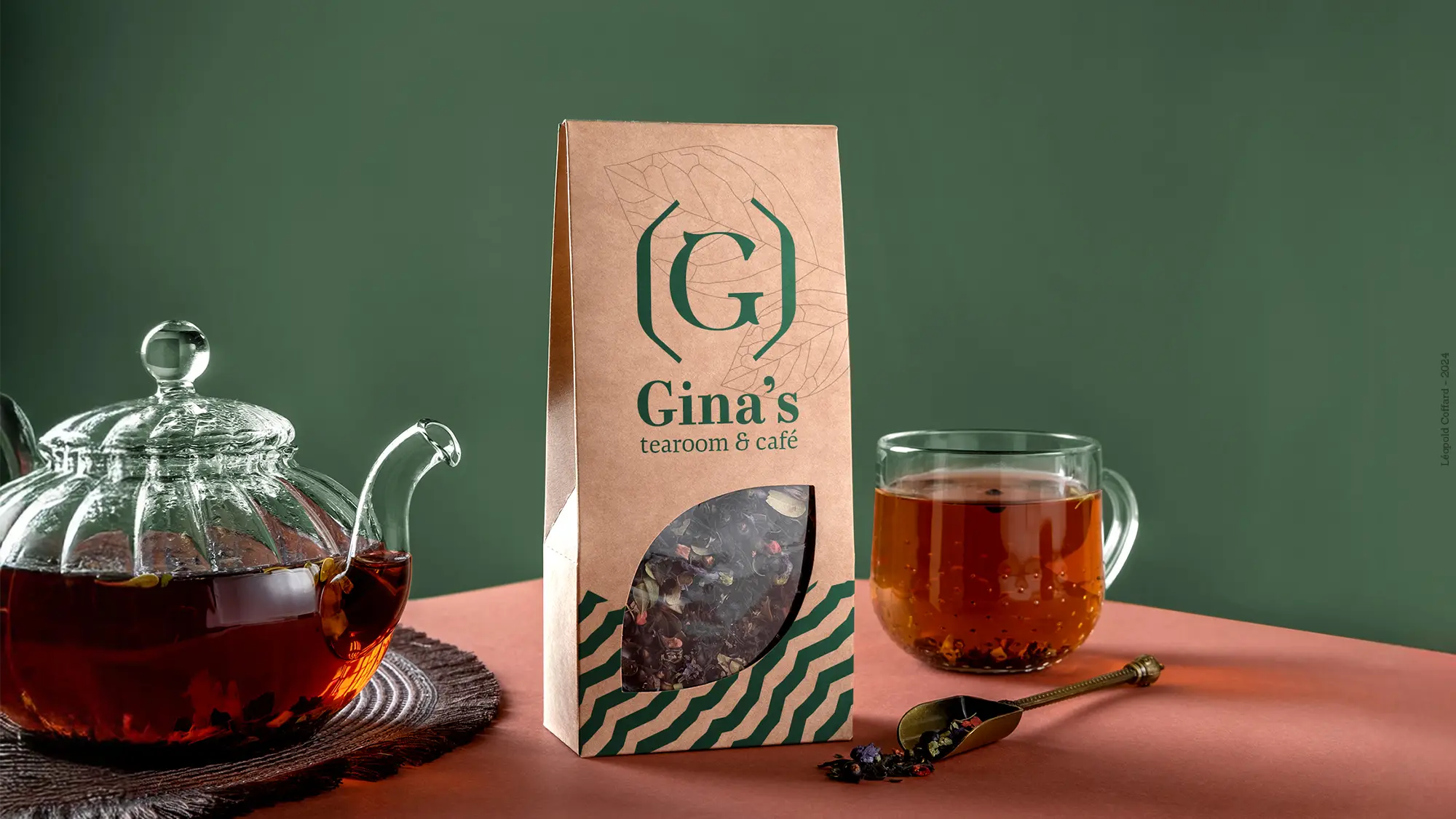 Glass teapot with brewed tea, brown paper package of Gina's tearoom & café tea leaves, and a glass cup of tea on a table with Gina’s tearoom & café logo on green background.