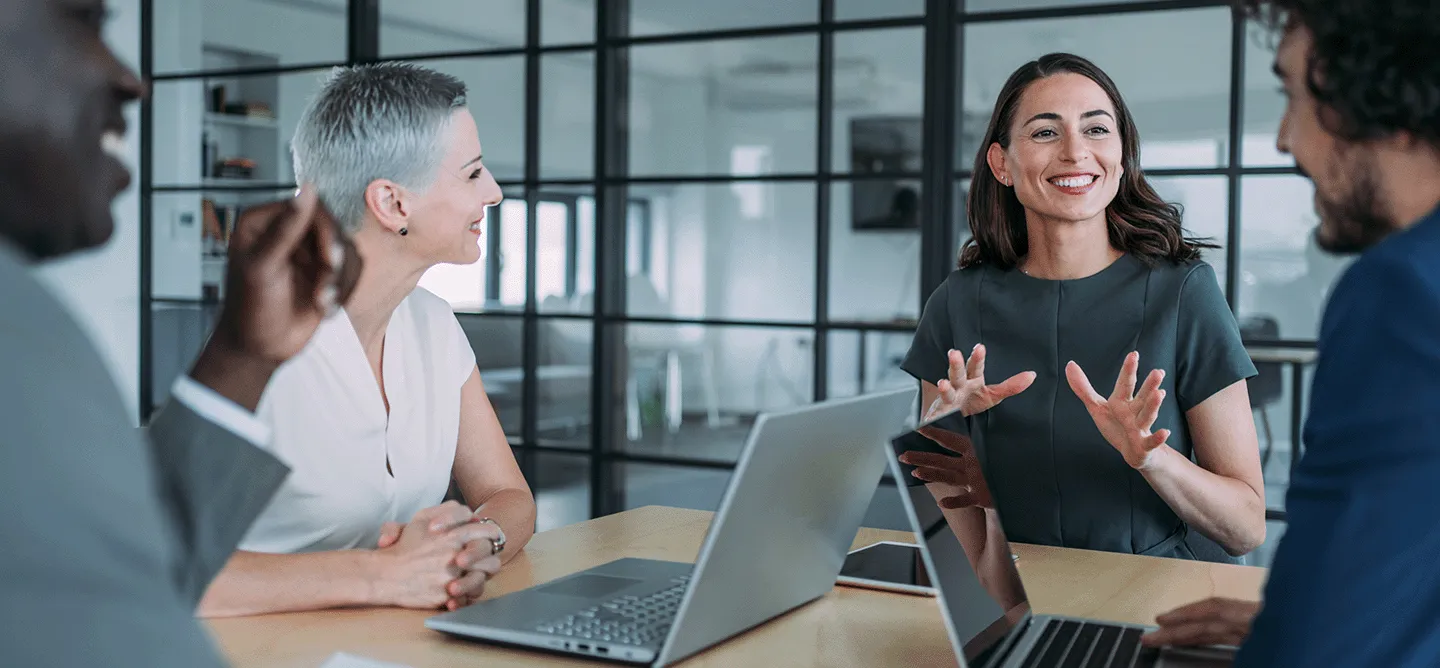 Mitarbeitende stehen vor ihren Laptops an einem Tisch und unterhalten sich