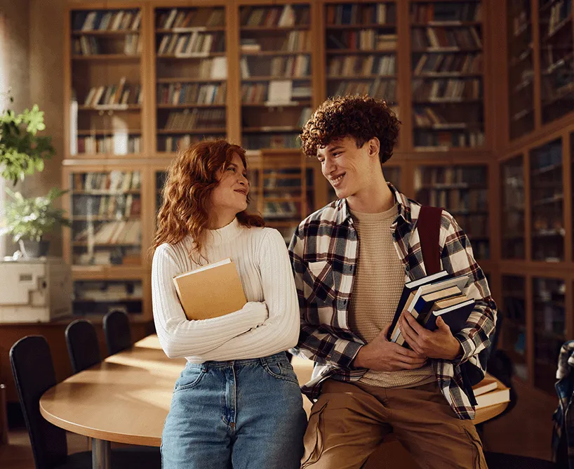 Zwei Studierende stehen in der Bibliothek gegen einen Tisch gelehnt und unterhalten sich