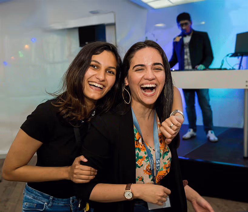 Zwei Frauen umarmen sich lachend auf einer Veranstaltung mit einem DJ im Hintergrund.