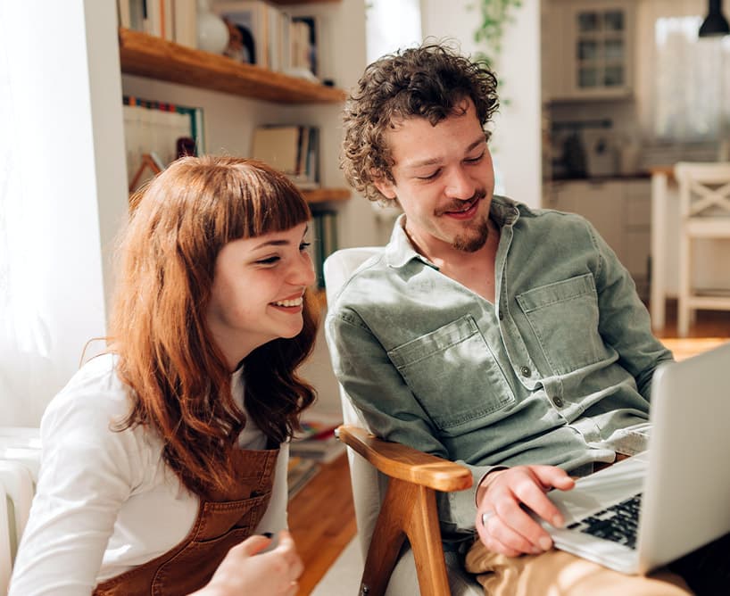 Junges Paar sitzt zu Hause zusammen und lächelt, während sie auf einen Laptop schauen.