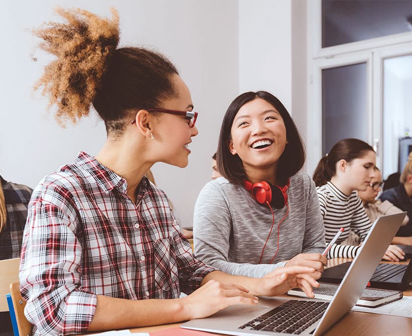 Zwei weibliche Studenten unterhalten sich lächelnd im Klassenzimmer, während sie an Laptops arbeiten.