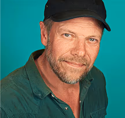 Middle-aged man with a beard wearing a black cap and green shirt against a teal background.