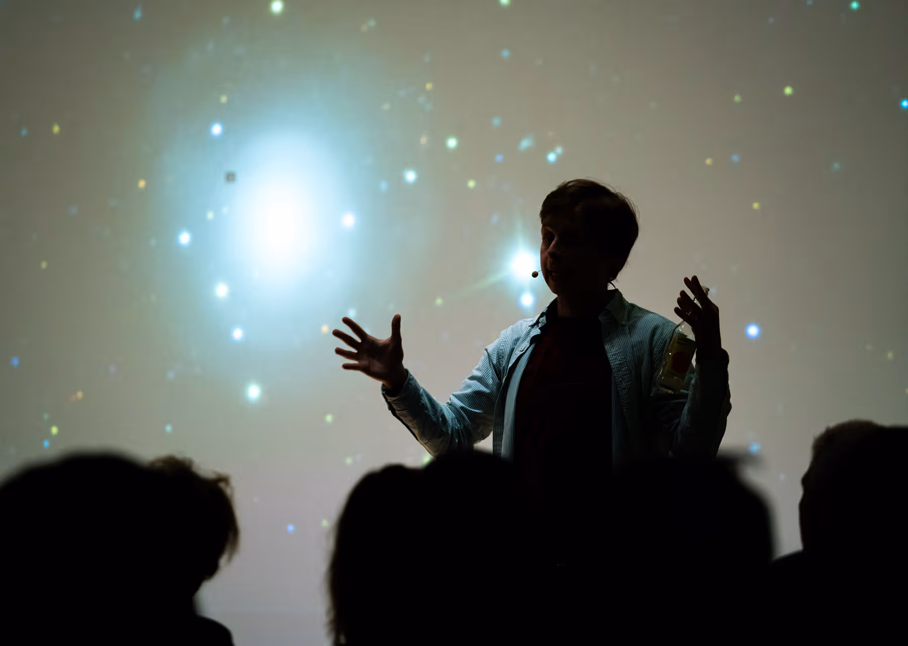 Silhouette of a person giving a presentation with a microphone and bottle, in front of a starry background.
