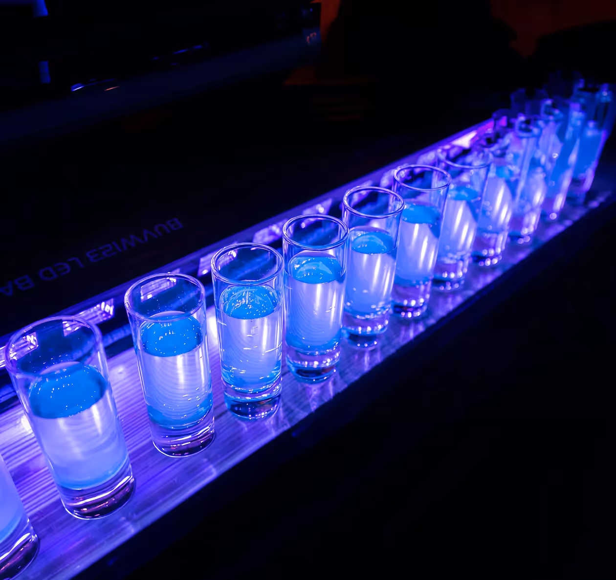 Row of shot glasses filled with a blue glowing liquid on a lit bar counter in a dark setting.