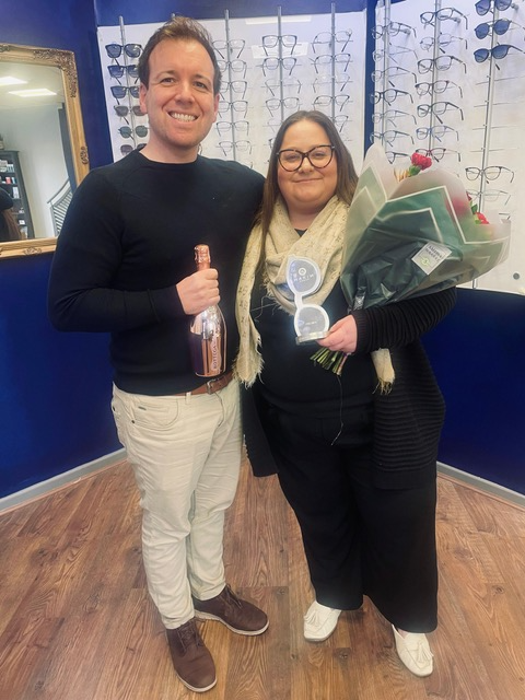 Two people smiling in eyeglass store, holding bottle and flowers