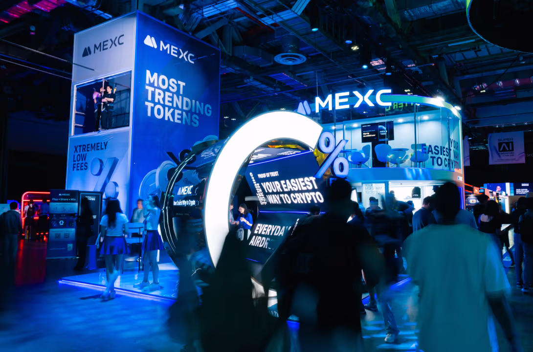 MEXC exhibition booth with bright blue lighting, featuring a large illuminated percentage symbol and promotional text about trending tokens and low fees.