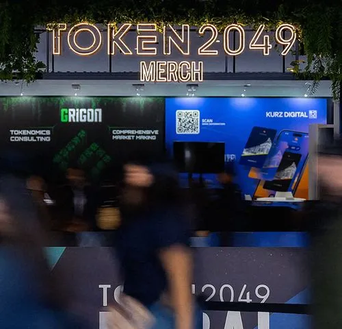 TOKEN2049 Dubai merchandise booth with illuminated sign and attendees browsing