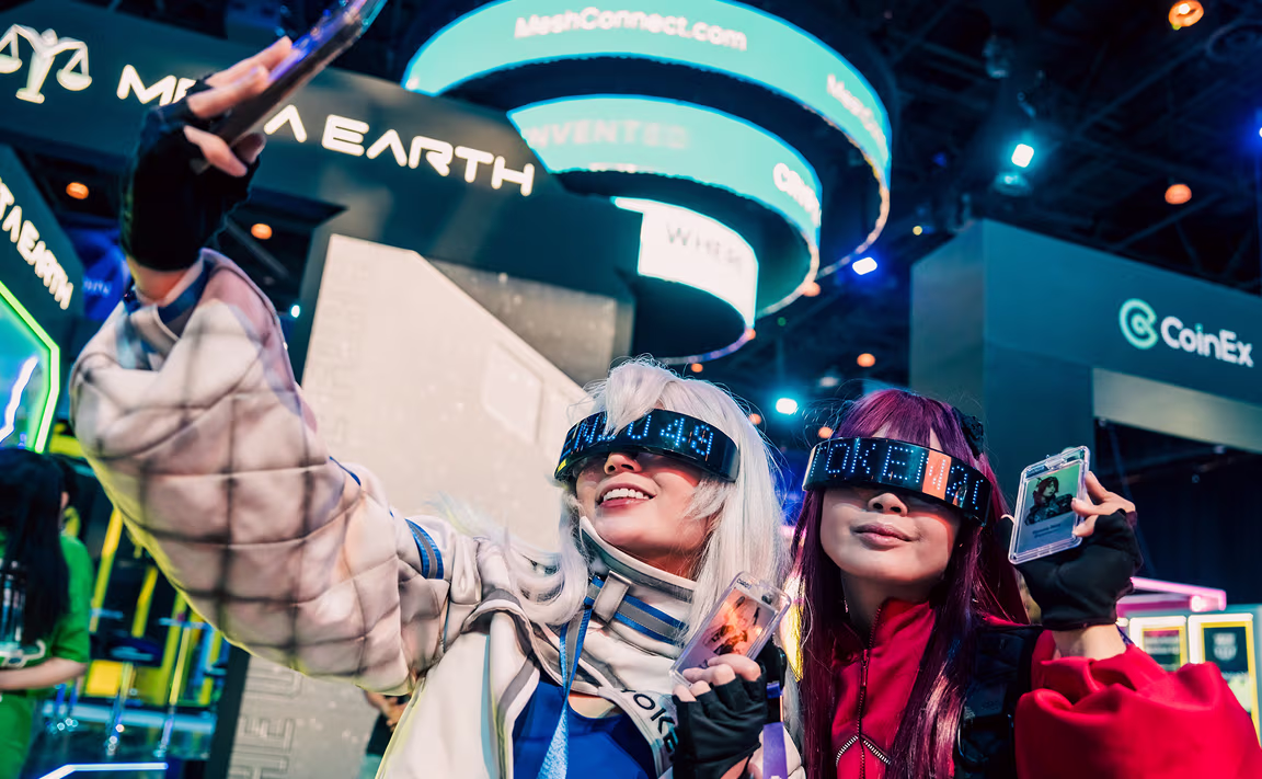 Two women wearing futuristic LED glasses and colorful costumes taking a selfie at a tech or gaming convention.