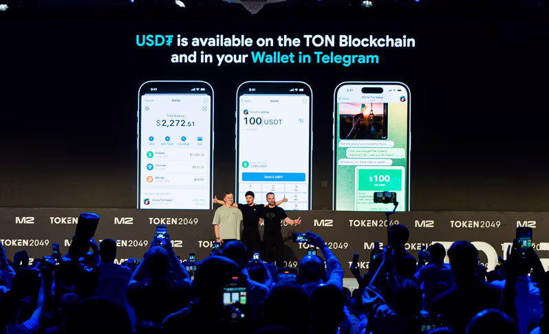 Three presenters on stage in front of a screen showing USD₮ available on TON Blockchain and Wallet in Telegram with app screenshots and an audience in foreground.