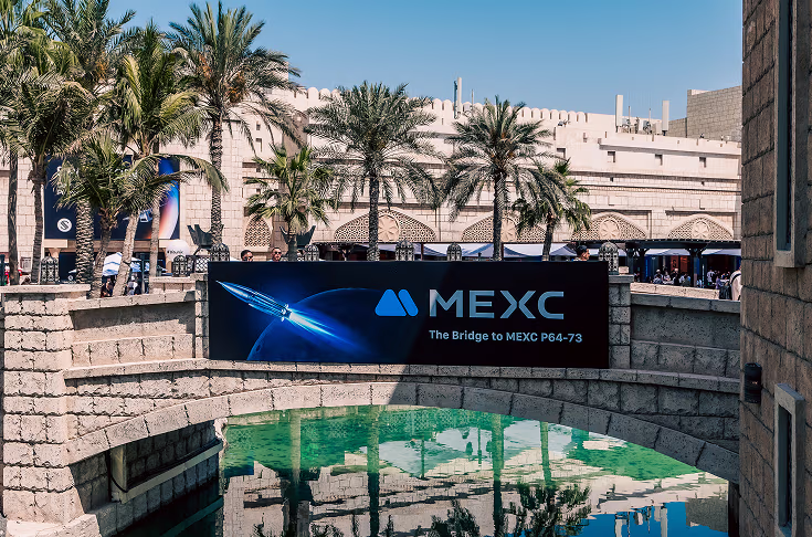 Stone bridge over green water with a MEXC banner reading 'The Bridge to MEXC P64-73,' palm trees, and a historic stone building in the background under a clear blue sky.