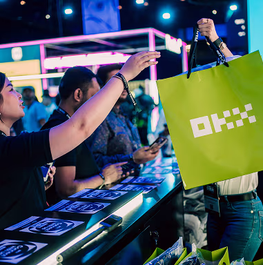A woman reaches out to receive a green shopping bag with a white logo from another person at a table in a busy, colorful indoor setting.