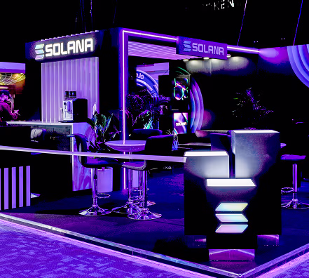 Exhibition booth with Solana branding illuminated in purple and blue lighting, featuring seating and modern decor.