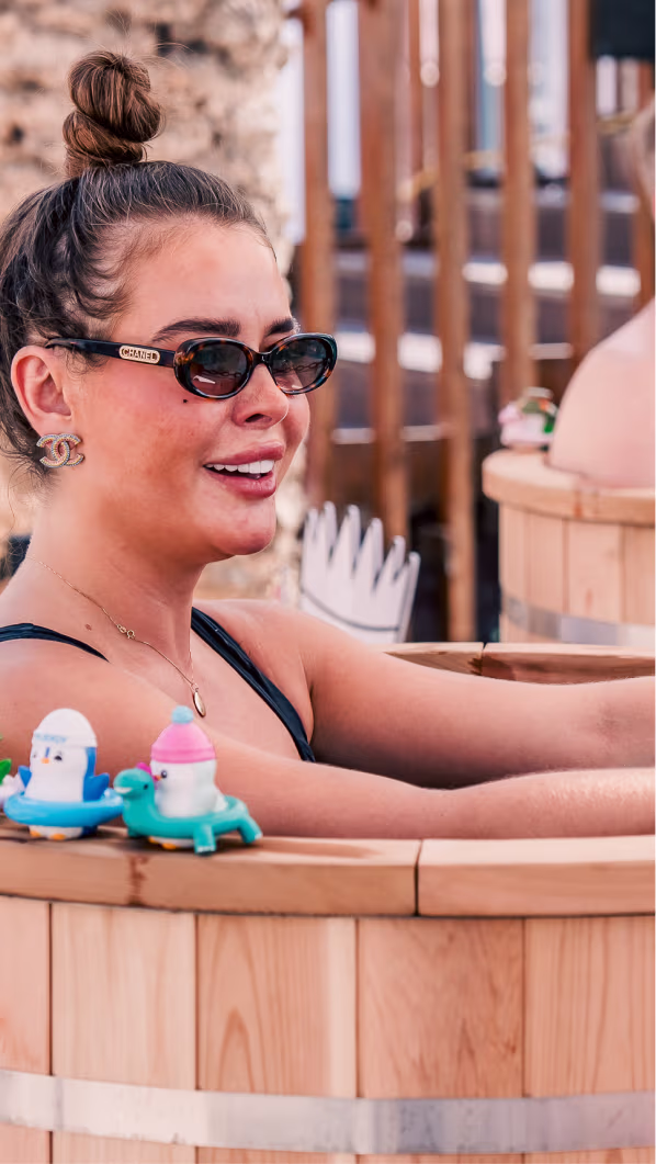 Smiling woman with sunglasses and a top bun relaxing in a wooden hot tub with small colorful toy figures on the edge.