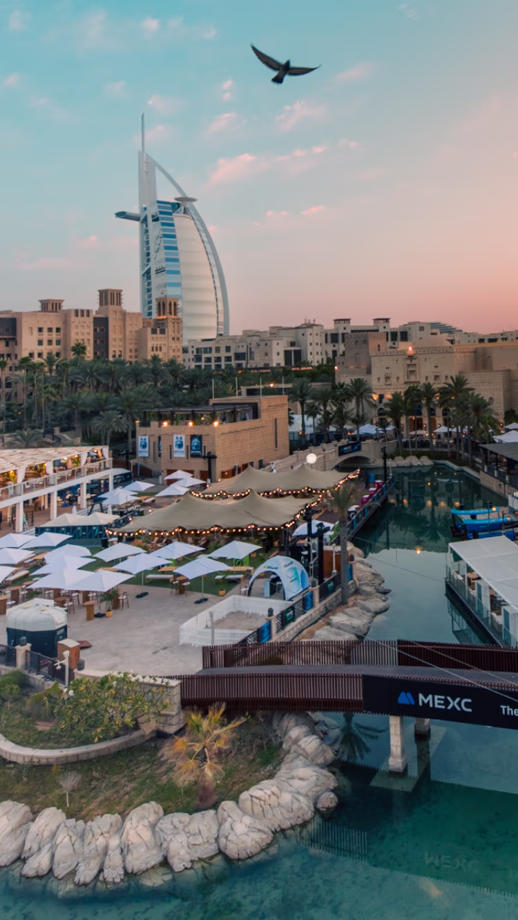 View of the Burj Al Arab hotel and surrounding buildings near a marina with outdoor seating and umbrellas at sunset.
