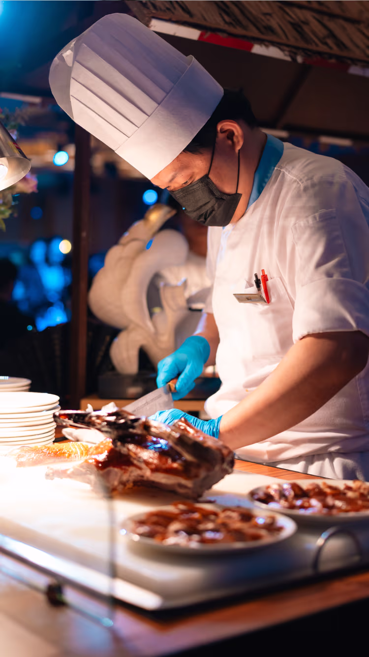 Chef wearing a white hat, face mask, and gloves slicing roasted meat on a cutting board in a restaurant kitchen.