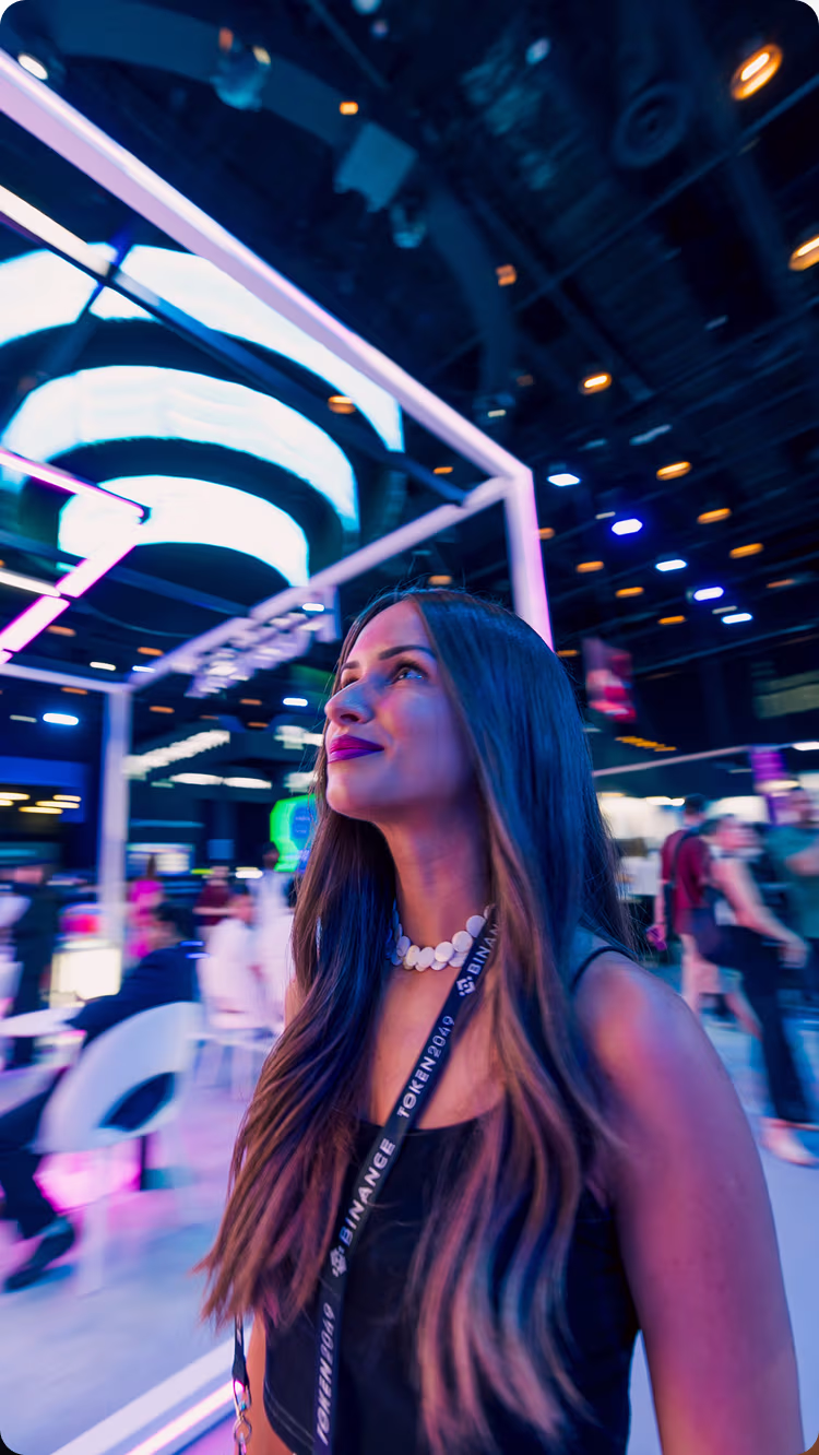 Young woman with long hair wearing a Binance lanyard and white necklace in a vibrant indoor event space with neon lighting.
