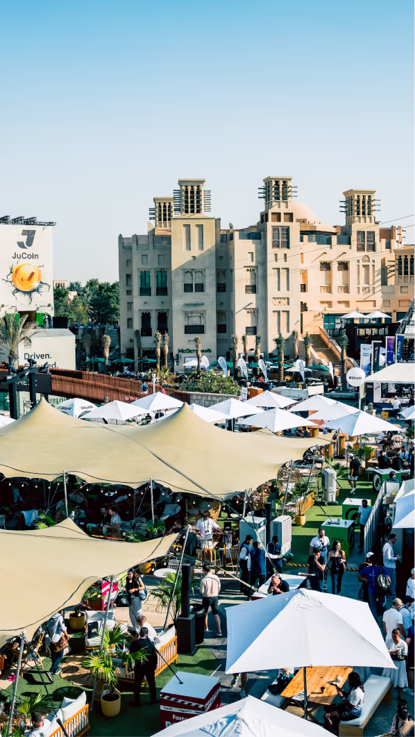 Outdoor event with people under large beige and white canopies and umbrellas, in front of a traditional beige building with wind towers.