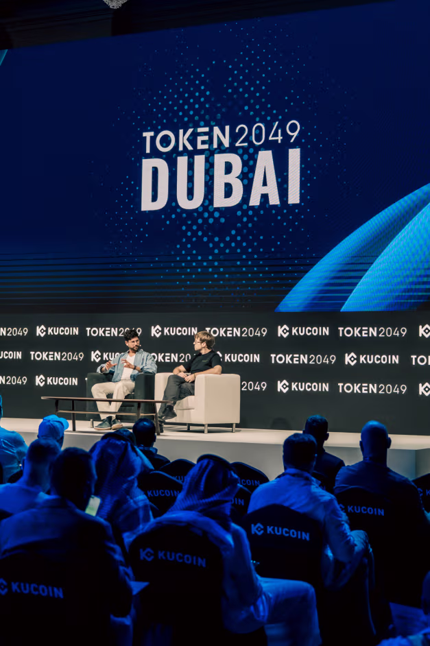 Two men seated on stage in armchairs having a discussion at TOKEN 2049 Dubai event with an audience wearing KuCoin shirts.