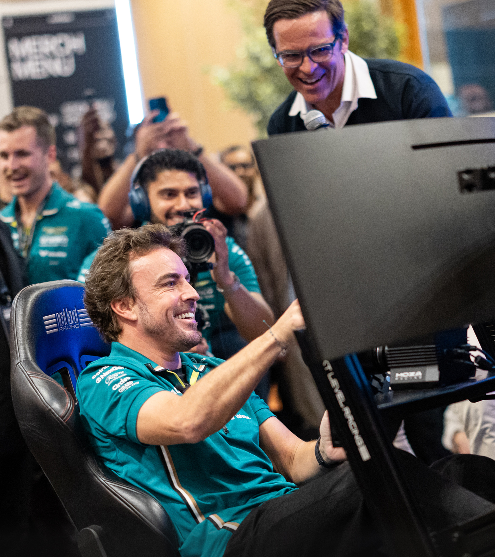 Smiling man in teal racing shirt sits in a racing simulator seat while others watch and take photos.