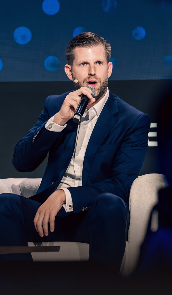 Man with microphone speaking while seated on a white chair against a dark background with blue light spots.