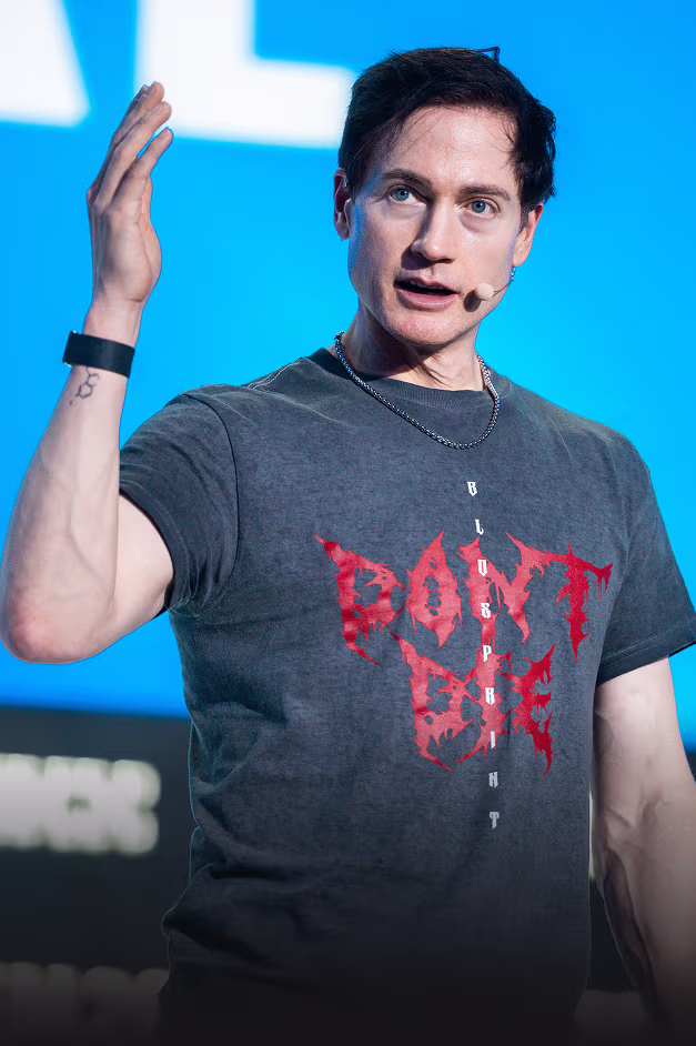 A man wearing a black t-shirt with red text 'DON'T BE' and a chain necklace speaking with a microphone headset against a blue background.